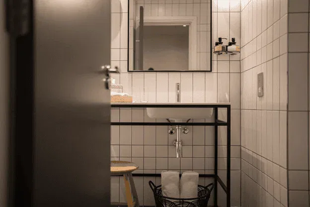 Ein modernes Badezimmer mit weiß gefliesten Wänden, einem rechteckigen Spiegel über einem weißen Waschbecken, einem Holzhocker auf der linken Seite und einem schwarzen Korb mit gerollten weißen Handtüchern darunter - die perfekte Inspiration für Ihre Altbau Badsanierung in Frankfurt.