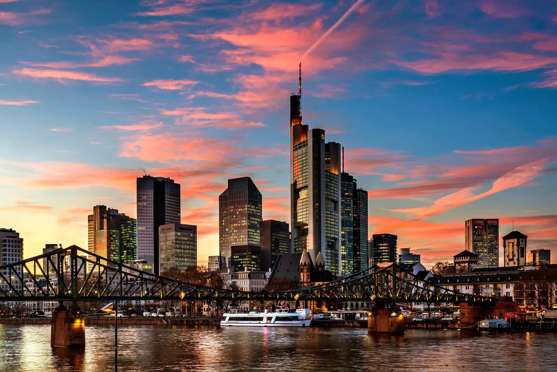 Eine Stadtsilhouette mit modernen Wolkenkratzern bei Sonnenuntergang, mit einer Brücke über einen Fluss im Vordergrund. Der leuchtende Himmel und die bunten Wolken schaffen eine Atmosphäre, die an einen automatisch gespeicherten Entwurf erinnert, der sich im Wasser spiegelt.