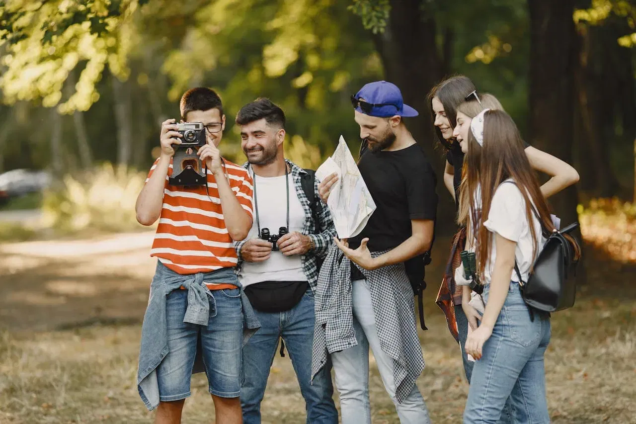 Eine Gruppe von fünf jungen Erwachsenen steht im Freien in einem Park oder Wald, stressfrei ins Umland unterwegs. Eine Person macht ein Foto, eine andere hält eine Karte in der Hand. Alle lächeln und genießen ihren gemeinsamen Ausflug.