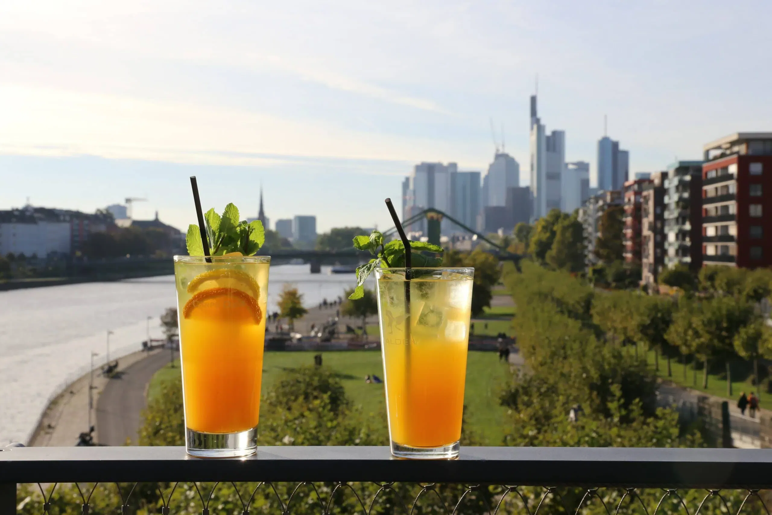 Zwei hohe Gläser mit orangefarbenen Cocktails mit Eis, Strohhalm und garniert mit Minze und Orangenscheiben stehen auf einem Geländer mit Blick auf einen malerischen städtischen Park am Flussufer. Im Hintergrund ist die Skyline des modernen Frankfurts mit seinen Wolkenkratzern, dem Fluss und Grünflächen zu sehen, was eine lebendige interaktive Szene erzeugt.