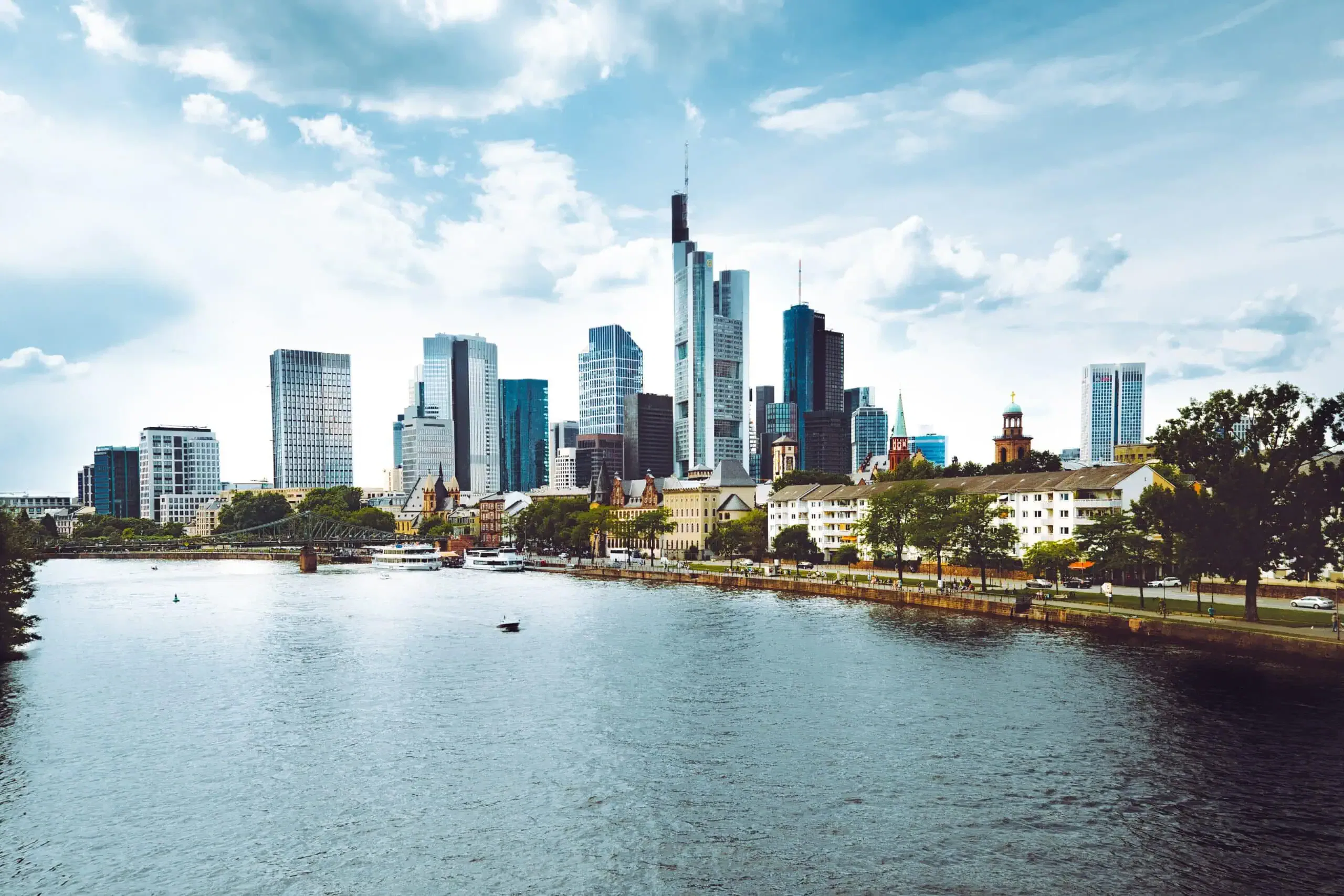 Panoramablick auf die Skyline von Frankfurt mit modernen Wolkenkratzern und Gebäuden entlang einer Uferpromenade. Der Fluss im Vordergrund spiegelt die Skyline wider, während der teilweise bewölkte Himmel einen dynamischen Hintergrund für diese Stadtlandschaft bietet. Interaktive Bäume säumen das Flussufer.
