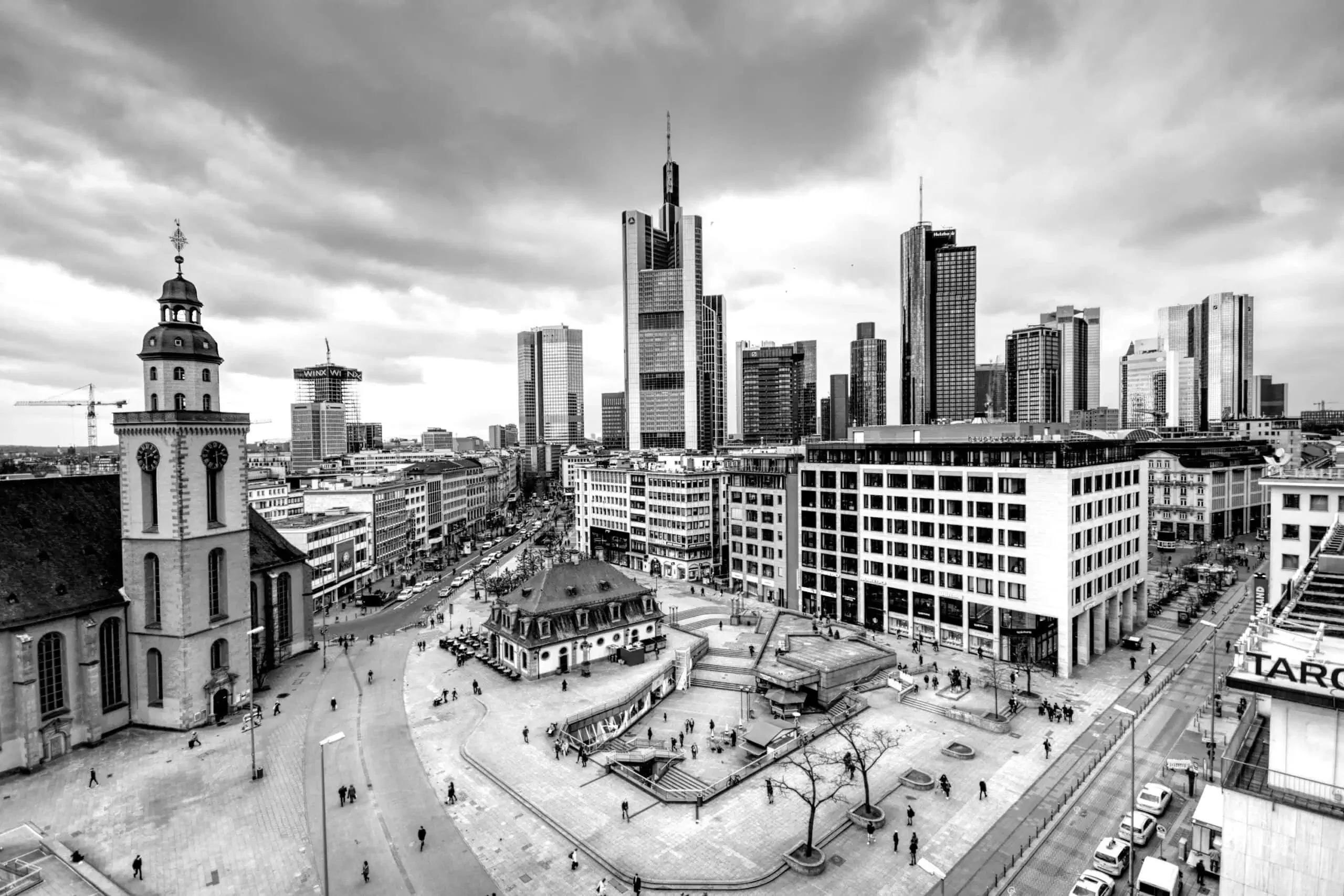 Schwarz-weiße Luftaufnahme von Frankfurt, Deutschland, zeigt eine Mischung aus modernen Wolkenkratzern und historischen Gebäuden. Die Skyline ist unter einem bewölkten Himmel zu sehen, mit Fußgängern, die sich um einen markanten Platz und eine Kirche im Vordergrund verteilen. Interaktive Elemente könnten diese Szene für den Betrachter zum Leben erwecken.