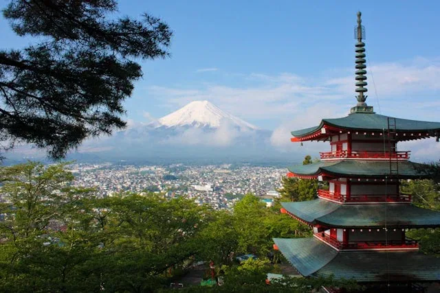 Eine rot-grüne Pagode steht inmitten von Bäumen, darunter eine Stadt und der Berg Fuji im Hintergrund, teilweise in Wolken gehüllt unter einem klaren blauen Himmel - die perfekte Inspiration für Ihre nächste Internationale Gruppenreise nach Japan.