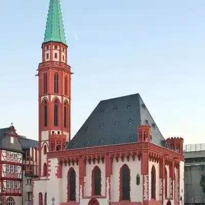 Eine historische Kirche mit einem hohen, spitzen grünen Turm und einer rot-weißen Außenfassade steht unter klarem Himmel. Die Kirche verfügt über Bogenfenster und aufwendige Verzierungen an den Wänden, was sie zu einer der herausragenden Sehenswürdigkeiten macht. In der Nähe ist ein traditionelles Fachwerkgebäude teilweise sichtbar.