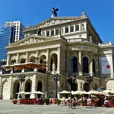 Das Bild zeigt ein großes historisches Gebäude mit neoklassizistischer Architektur, die Alte Oper in Frankfurt. Das Gebäude verfügt über verzierte Säulen, Skulpturen und eine Statue auf dem Dach. Man sieht Sitzgelegenheiten im Freien mit Sonnenschirmen und im Hintergrund moderne Gebäude, was es zu einer der bemerkenswerten Sehenswürdigkeiten der Stadt macht.