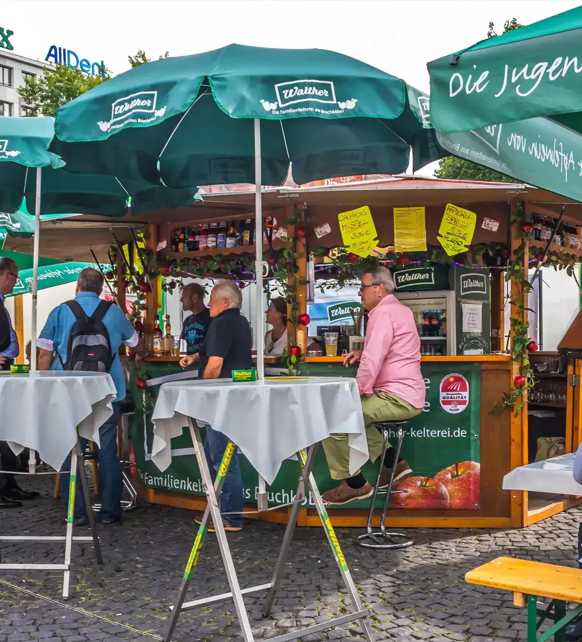 An einem belebten Stand im Freien beim Apfelwein-Festival stehen Menschen um hohe, mit weißen Tischdecken bedeckte Tische herum und genießen Getränke. Der Stand verfügt über grüne Schirme mit dem „Weltheis“-Logo und verschiedene Schilder und Dekorationen. Die Atmosphäre wirkt lebhaft und gesellig.