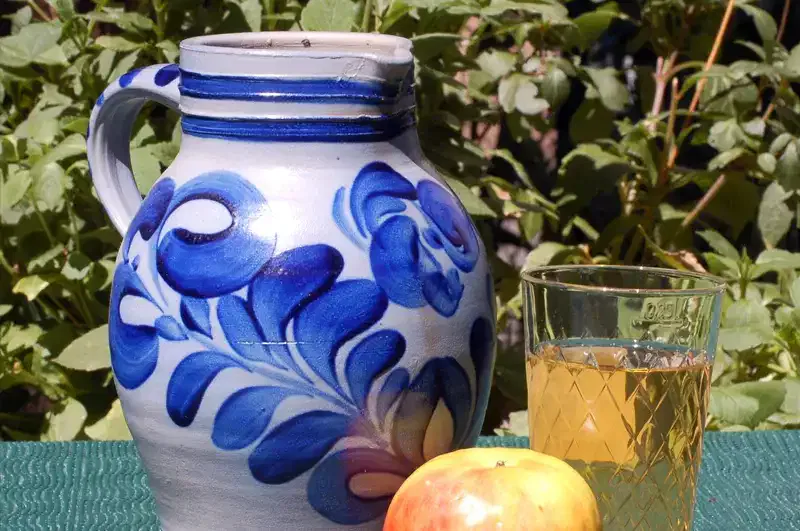 Ein Keramikkrug mit blauem Blumenmuster steht neben einem Glas Apfelsaft und einem rot-gelben Apfel auf einer grünen Tischdecke. Im Hintergrund sind grüne Blattpflanzen zu sehen, die die perfekte Kulisse für ein Apfelweinfest schaffen.