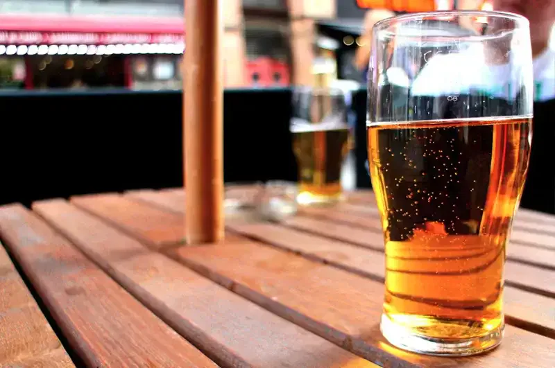Ein Pint bernsteinfarbenes Bier steht auf einem hölzernen Tisch im Freien. Im Hintergrund sind ein weiteres Bier und ein Aschenbecher zu sehen, zusammen mit einem roten Gebäude und verschwommenem Laub, was eine entspannte Atmosphäre beim Apfelweinfest suggeriert.