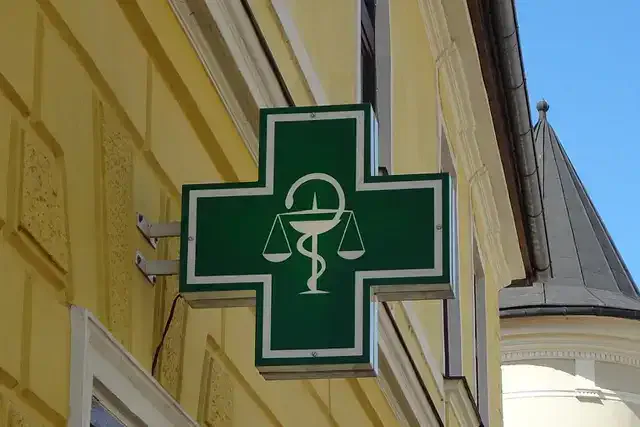 Ein grünes Kreuz mit dem weißen Symbol des Äskulapstabs und der Waage, das auf die Dr. Cech Apotheke hinweist, ist an der Ecke eines gelben Gebäudes mit architektonischen Details angebracht. Im Hintergrund ist vor einem klaren blauen Himmel die Turmspitze eines anderen Gebäudes zu sehen.