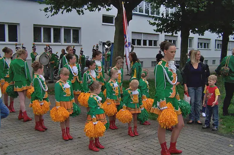 Eine Gruppe von Kindern in grünen Jacken, gelben Röcken und roten Stiefeln mit Pompons tritt bei der Bernemer Kerb-Parade auf. Sie stehen in Formation mit Erwachsenen und Zuschauern in der Nähe. Einige Leute sind leger gekleidet und im Hintergrund ist ein Gebäude zu sehen.