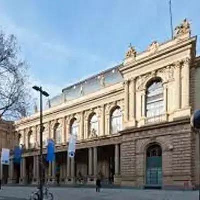 Das Bild zeigt ein großes historisches Gebäude mit klassischer Architektur, hohen Bogenfenstern und kunstvollem Mauerwerk. Vor der Sehenswürdigkeit hängen mehrere blau-weiße Flaggen an Masten, und im Vordergrund sind einige Menschen und Fahrräder zu sehen.