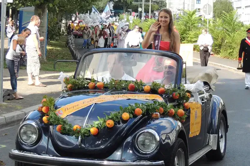 Eine Frau in einem roten Kleid steht lächelnd und winkend in einem blauen, mit Blumen geschmückten VW Käfer Cabrio. Das Auto ist Teil der Bernemer Kerb-Parade, bei der Zuschauer und Teilnehmer in verschiedenen Kostümen den Hintergrund mit lebhafter Feststimmung füllen.