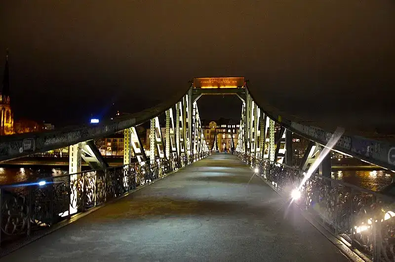 Eine gut beleuchtete Fußgängerbrücke mit Metallgeländer, bekannt als Eiserner Steg, erstreckt sich nachts über einen Fluss. Im Hintergrund sind Gebäude und ein Kirchturm zu sehen, beleuchtet von den Lichtern der Stadt. Der Himmel ist dunkel und bildet einen auffälligen Kontrast zur hell erleuchteten Brücke.
