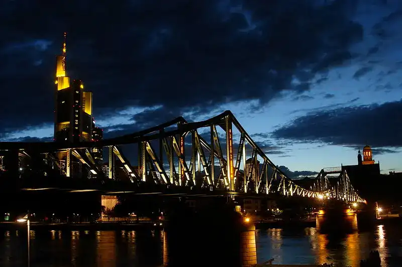 Nachtansicht einer beleuchteten Brücke, die sich über einen Fluss spannt, wobei sich die Lichter der Stadt im Wasser spiegeln. Der Himmel ist dunkel mit vereinzelten Wolken, und die Gebäude auf beiden Seiten der Brücke sind ebenfalls beleuchtet, was eine ruhige Stadtlandschaft schafft.