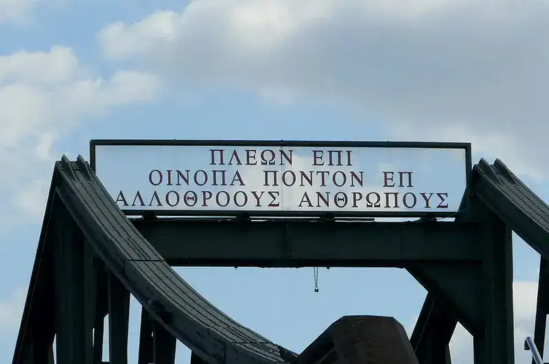 Eine Metallbogenbrücke, die an den Eisernen Steg erinnert, ist oben mit einem Schild versehen, auf dem ein griechischer Text in roten Buchstaben auf weißem Hintergrund zu sehen ist. Der Himmel dient als malerische Kulisse für die Worte: „ΠΛΕΩΝ ΕΠΙ ΟΙΝΟΠΑ ΠΟΝΤΟΝ ΕΠΙ ΑΛΛΟΘΡΟΟΥΣ ΑΝΘΡΩΠ