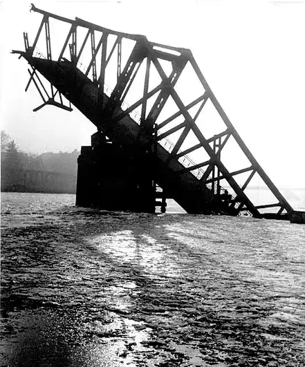 Eine kaputte und teilweise eingestürzte Stahlkonstruktion, die dem Eisernen Steg ähnelt, steht über einem Gewässer. Die zerklüfteten Überreste der beschädigten Brücke heben sich als Silhouette vor einem hellen, dunstigen Himmel ab. Das Wasser darunter reflektiert das Licht und im Hintergrund sind Bäume zu sehen.