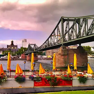 Eine malerische Aussicht auf einen Fluss, über den eine Stahlbrücke führt. Darunter befindet sich ein Café am Flussufer, das mit leuchtend gelben Sonnenschirmen und bunten Blumenkästen geschmückt ist. Im Hintergrund sind Gebäude und ein teilweise bewölkter Himmel zu sehen – wahrlich eine der bezaubernden Sehenswürdigkeiten der Gegend.