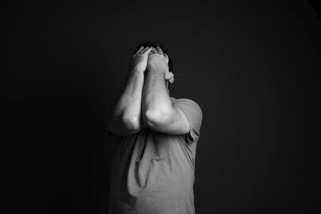 Eine Person, die ein T-Shirt trägt, steht vor einem dunklen Hintergrund und bedeckt ihr Gesicht mit beiden Händen und angewinkelten Ellbogen, was auf organisierten Stress oder Bedrängnis hindeutet - vielleicht spiegelt es Abschiede oder emotionale Belastungen wider, denen Angehörige ausgesetzt sind. Das Bild ist in schwarz-weiß gehalten.