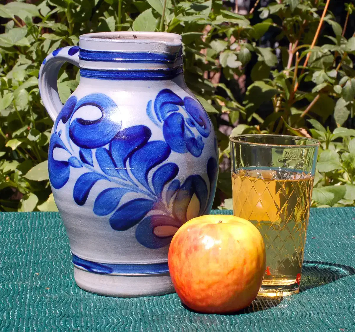 Ein blau-weißer Keramikkrug mit Blumenmuster steht im Freien auf einer grünen Tischdecke. Daneben stehen ein Glas Apfelwein und ein frischer rot-gelber Apfel. Im Hintergrund sind grüne Blätter zu sehen.