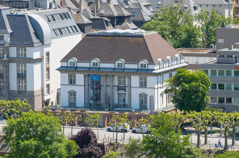 frankfurt-deutsches-architektur-museum Ein großes, historisches Gebäude mit Satteldach und Säulen am Eingang, gelegen in einer städtischen Umgebung inmitten umliegender Gebäude und üppiger Bäume. Die am malerischen Museumsufer gelegene Baumreihe und die geparkten Autos tragen zum charmanten Straßenbild bei.