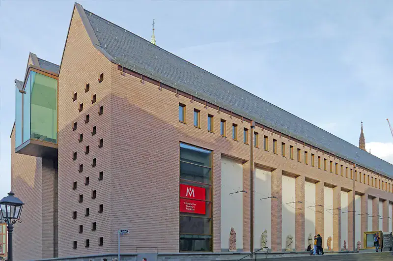 frankfurt-historisches-museum Außenansicht des Historischen Museums der Pfalz in Speyer, Deutschland. Die moderne Fassade ist eine Mischung aus traditionellem und zeitgenössischem Stil mit einem großen, schrägen Dach und einem bemerkenswerten Glasvorsprung, der an die Architektur am Museumsufer erinnert. Ein rotes Museumsbanner schmückt die Vorderwand.
