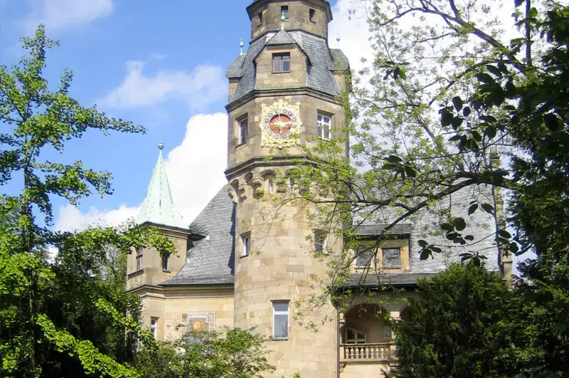 frankfurt-liebieghaus An einem klaren, sonnigen Tag steht inmitten üppiger Vegetation ein historisches Steinschloss mit einem hohen zentralen Turm, der mit einer großen Uhr geschmückt ist und mit grauen Schindeln gedeckt ist. Teilweise von Bäumen verdeckt, erinnert diese malerische Szene unter einem mit vereinzelten Wolken übersäten Himmel an das charmante Museumsuferviertel.