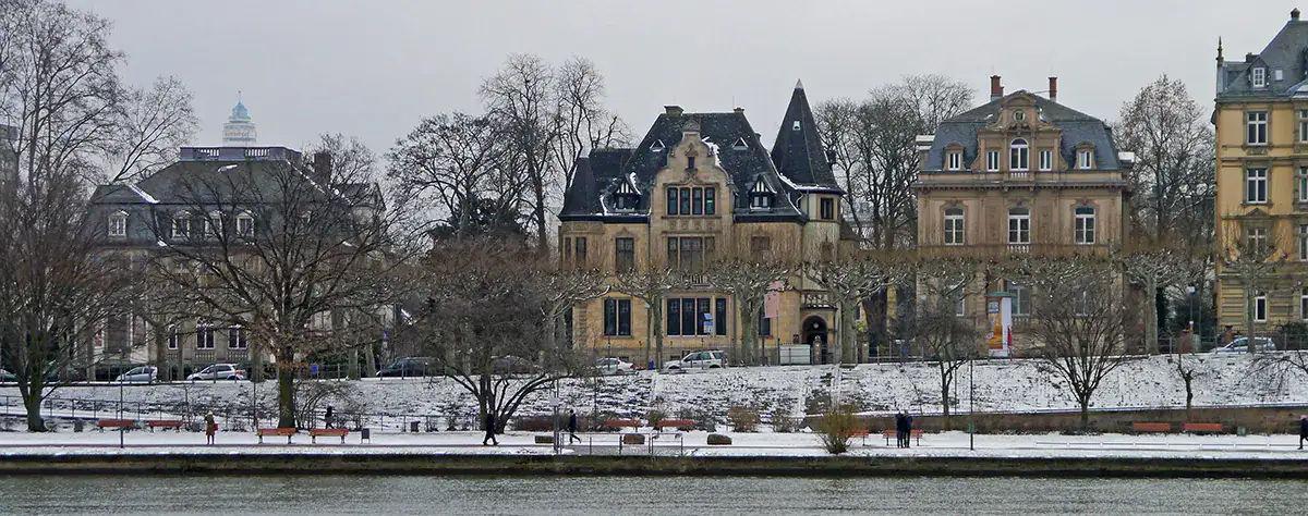 frankfurt-museum-der-weltkulturen Eine Winterlandschaft zeigt stattliche, historische Gebäude entlang der verschneiten Uferpromenade des Museumsufers. Kahle Bäume rahmen das Gebiet ein, und Bänke und Wege verlaufen durch den schneebedeckten Park. Das ruhige Wasser spiegelt den gedämpften, bedeckten Himmel wider, der typisch für einen kalten Wintertag ist.