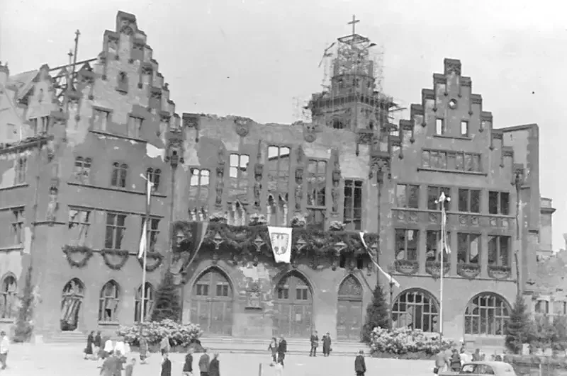 frankfurt-roemer-1947 Schwarzweißfoto eines großen, beschädigten Gebäudes mit Treppengiebeln, eingerüstetem Turm und gotischer Architektur, die an den Römer erinnert. Einige Fenster sind zerbrochen und Teile des Gebäudes scheinen restauriert zu werden. Im Vordergrund laufen Menschen.