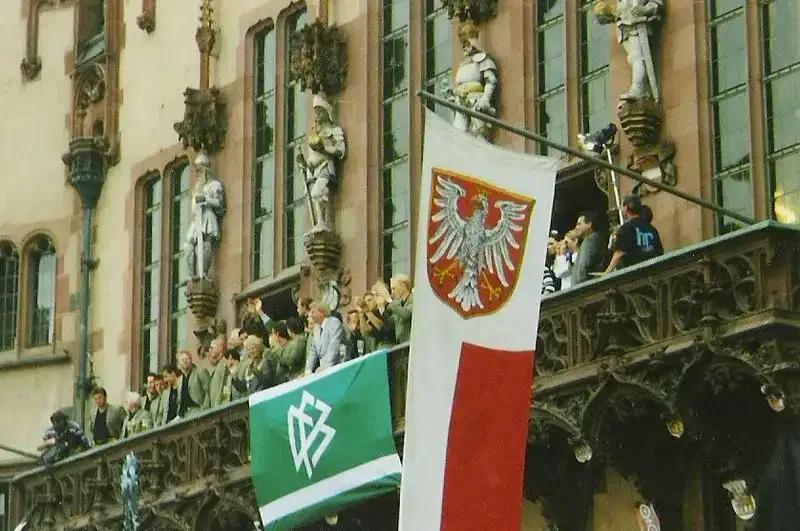frankfurt-roemer-balkon Eine Gruppe von Menschen steht auf einem mit Statuen geschmückten Balkon des historischen Römers. Davor hängen zwei Flaggen: eine mit einem weißen Adler auf rotem Grund und eine mit einem Symbol, das einer Raute ähnelt, in Weiß auf grünem Grund.