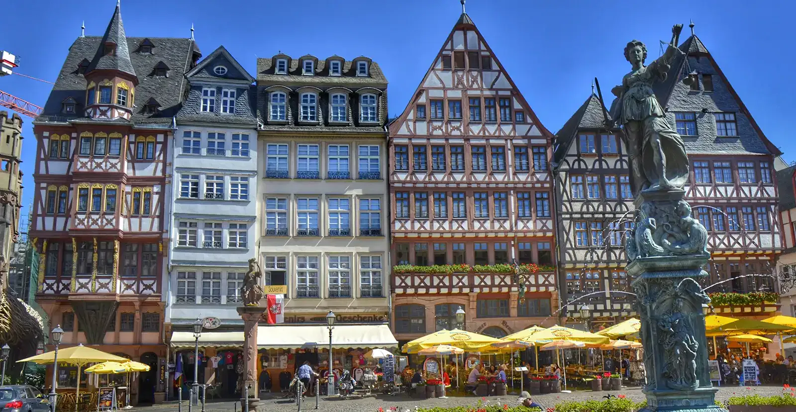 frankfurt-roemer-fassade Eine Reihe farbenfroher, kunstvoll verzierter Fachwerkhäuser säumt eine Kopfsteinpflasterstraße in einer europäischen Stadt. Davor wimmelt es in Straßencafés mit gelben Sonnenschirmen von Menschen. Eine dekorative Statue mit einer weiblichen Figur, die an den Römer erinnert, steht prominent im Vordergrund. Der Himmel ist klar und blau.