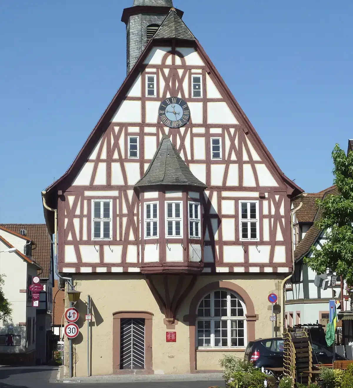 Ein Fachwerkhaus mit steilem, dreieckigem Dach und einem kleinen Türmchen mit einer Uhr steht im hellen Tageslicht von Bergen-Enkheim. Das Gebäude hat ein beiges Erdgeschoss mit einem gewölbten Eingang. Rundherum sind saubere Straßen, Verkehrsschilder und Grünflächen.