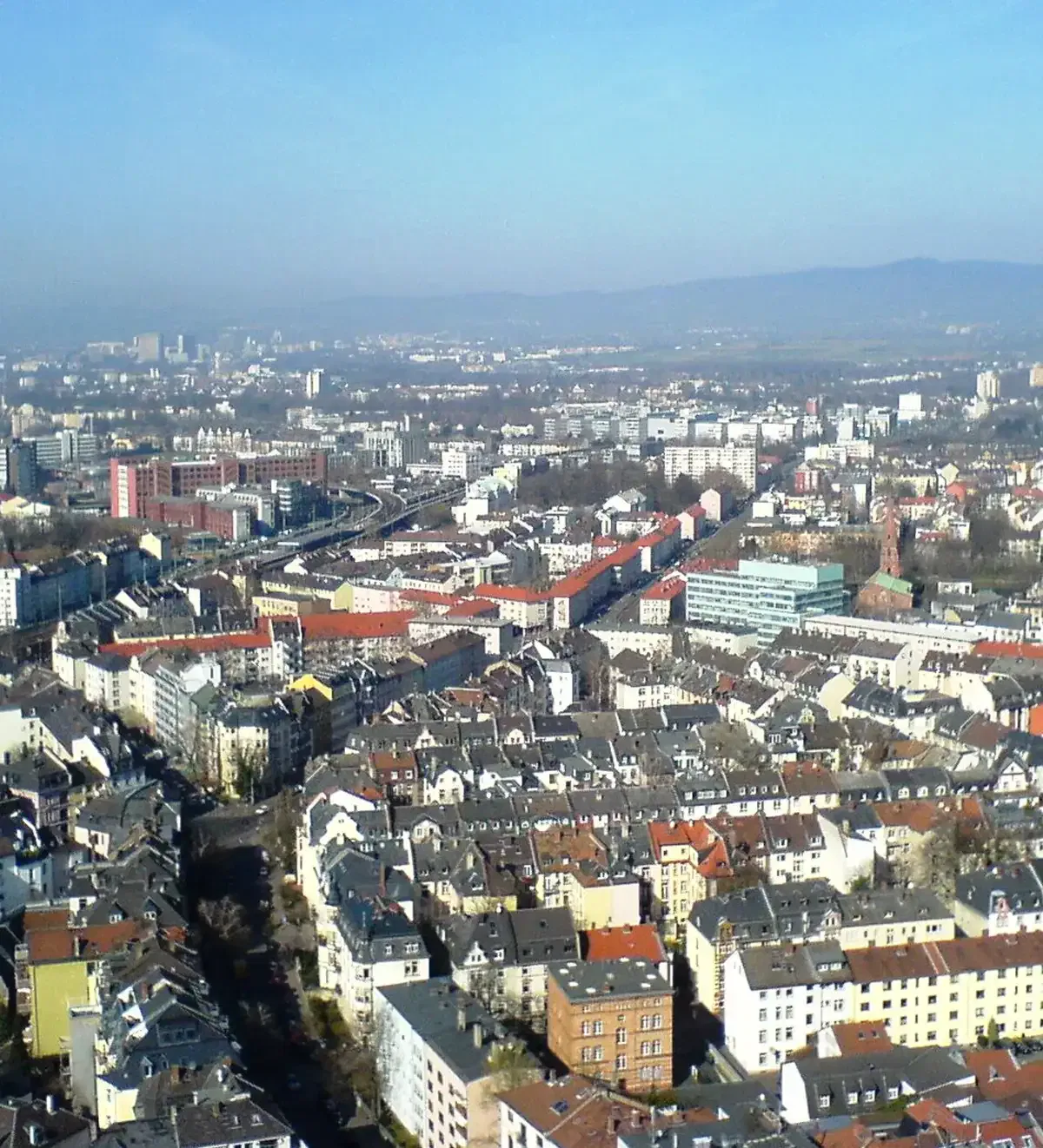 frankfurt-stadtteile-bockenheim Luftaufnahme der Stadtlandschaft von Bockenheim mit dicht gedrängten Gebäuden und einer Vielzahl von roten, gelben und grauen Dächern. Die Stadt ist mit Bäumen und breiten Straßen durchsetzt. Im Hintergrund sind Hügel und ein dunstiger blauer Himmel sichtbar.