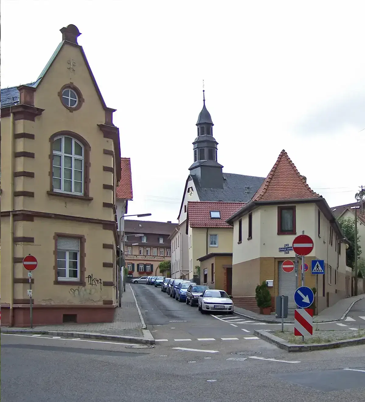 Eine Straßenszene in der malerischen Stadt Bonames zeigt schmale Straßen, gesäumt von alten Gebäuden mit schrägen Dächern. Autos parken an den Seiten und im Hintergrund erhebt sich majestätisch eine Kirche mit einem hohen Glockenturm. Verkehrsschilder und ein Fußgängerüberweg sind zu sehen.