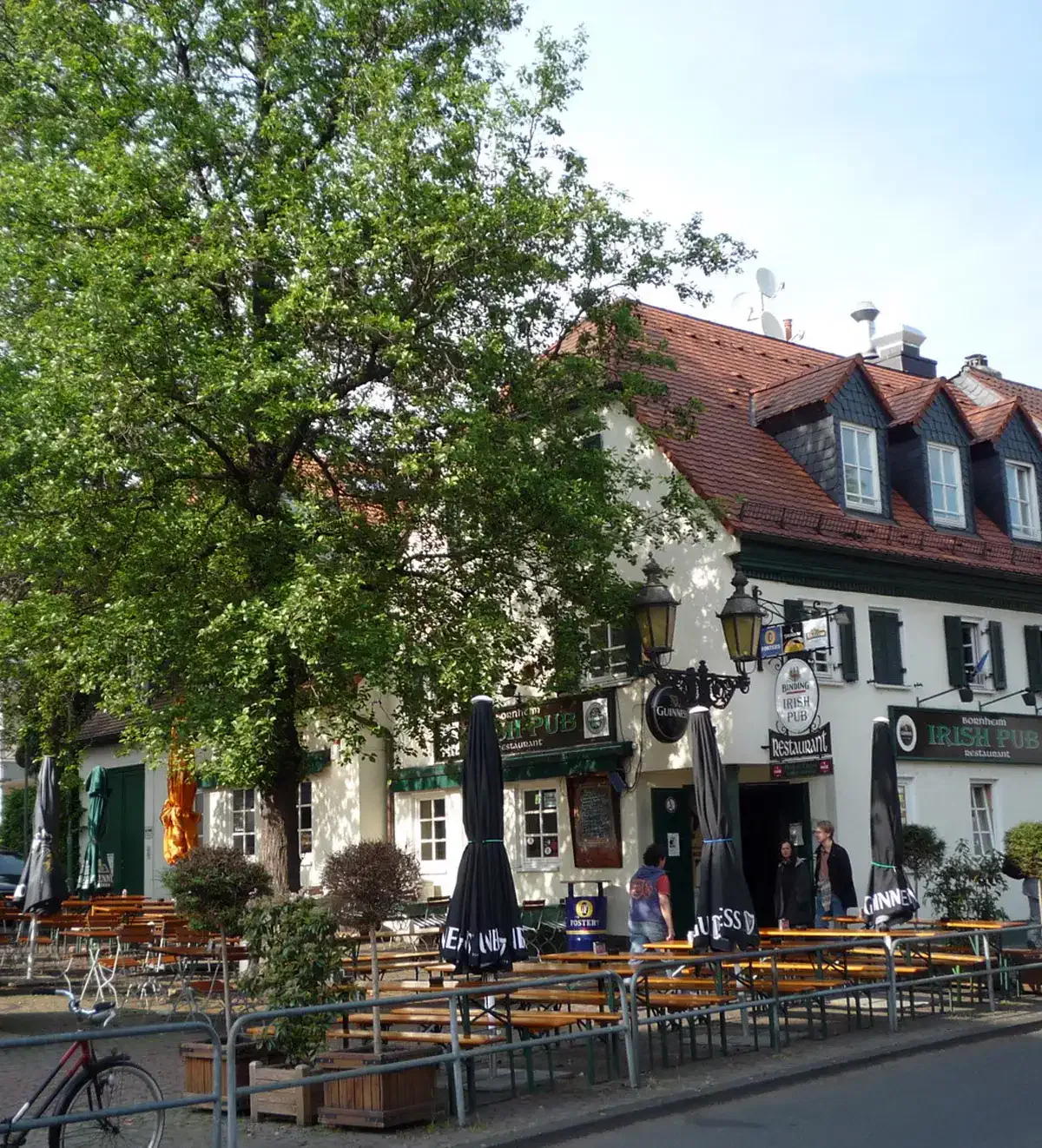 Ein gemütlicher Außensitzplatz mit Holztischen und Bänken unter Sonnenschirmen vor einem Irish Pub in Bornheim. Das Gebäude ist weiß mit einem roten Ziegeldach und ein großer Baum spendet Schatten. Die Leute sitzen und gehen vorbei, was eine entspannte Atmosphäre schafft.