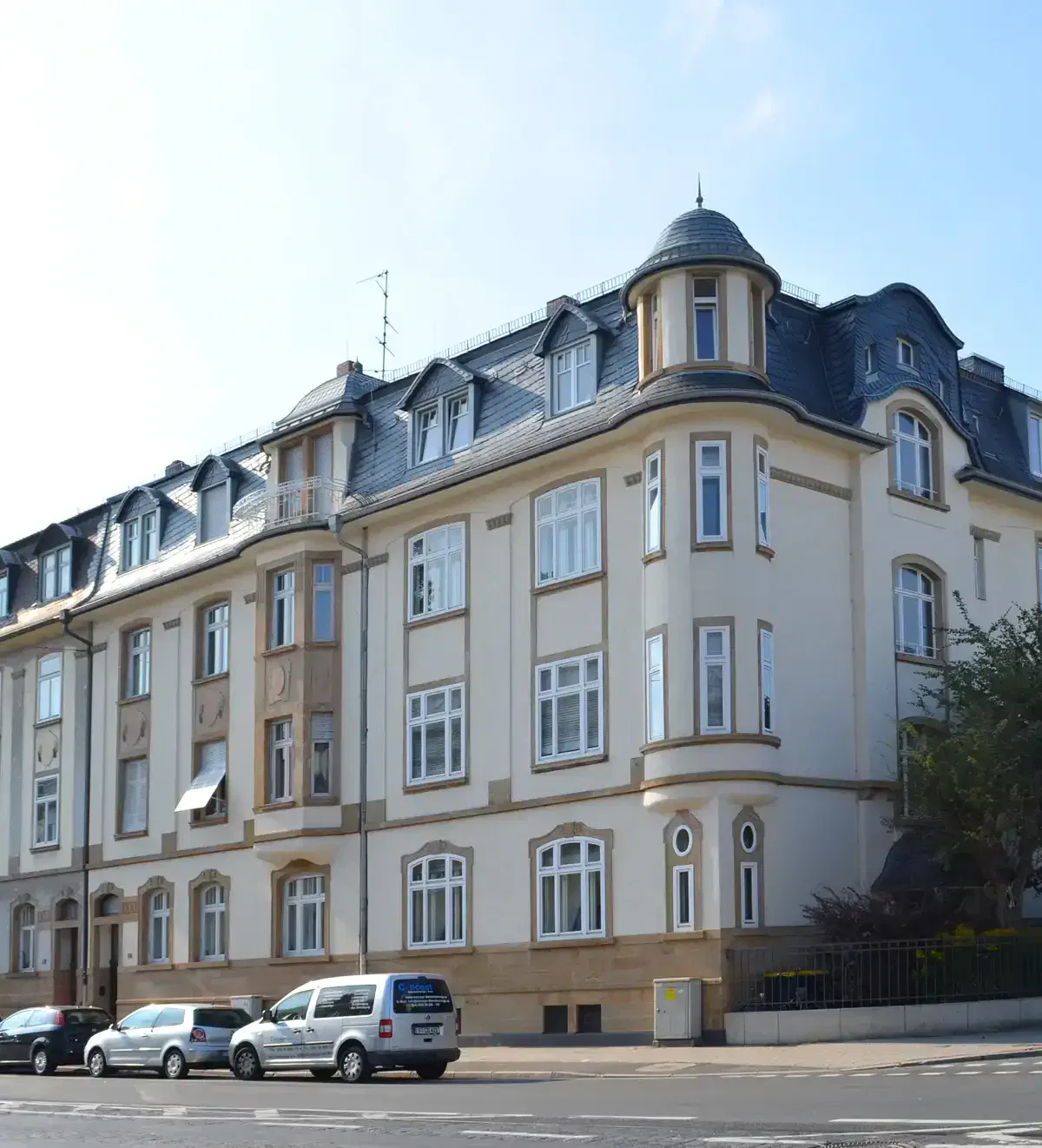 Ein großes, mehrstöckiges Gebäude mit cremefarbenen Wänden und einem Schieferdach ragt prominent in das Viertel Dornbusch. Das Gebäude verfügt über zahlreiche Fenster, zwei abgerundete Ecktürme mit konischen Dächern und eine Mischung aus rechteckigen und gewölbten Fenstern. Entlang der Straße davor sind mehrere Autos geparkt.