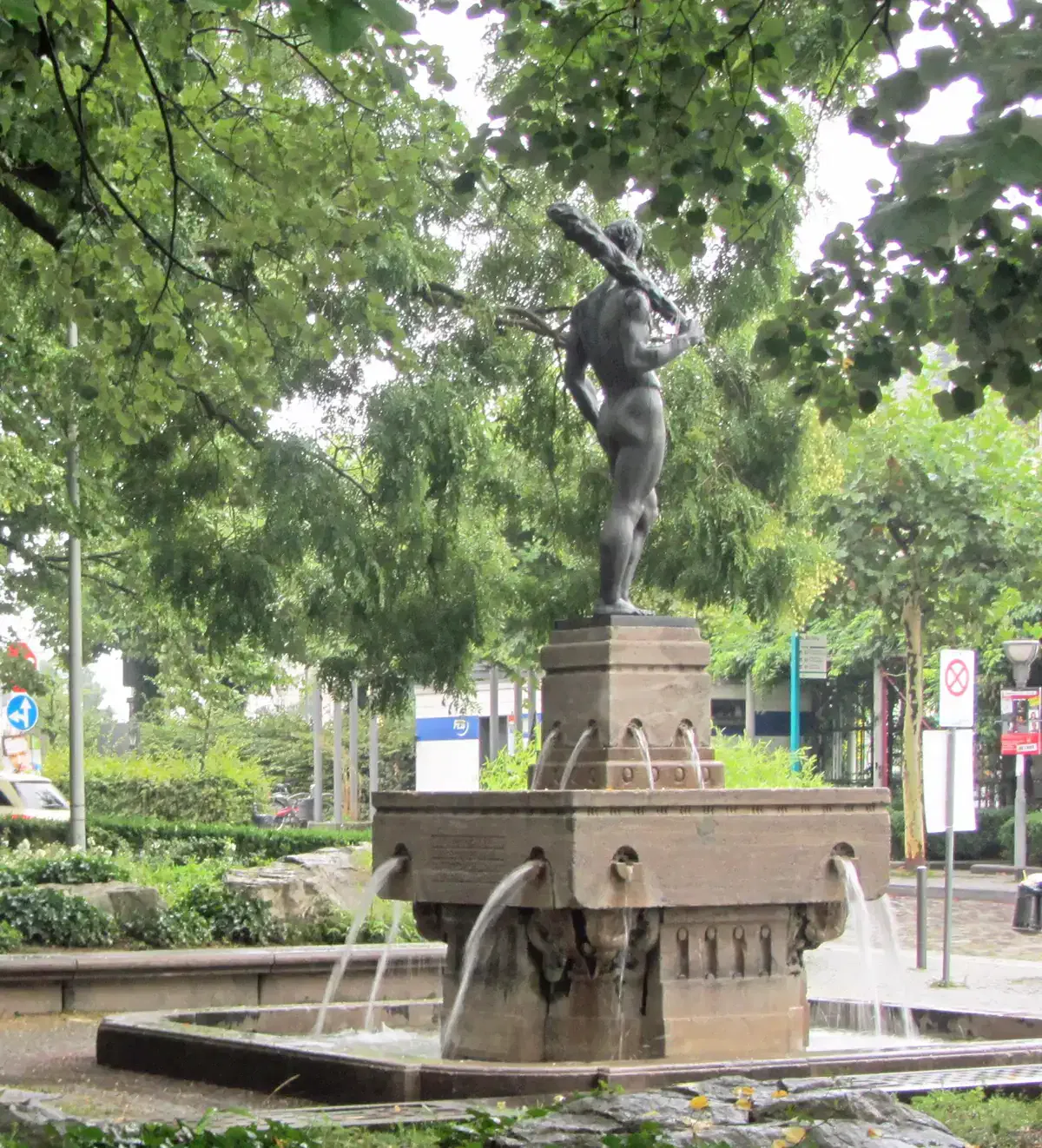 Die Statue einer Person mit einer Keule steht zentral auf einem erhöhten Sockel in einem Brunnen, aus dessen Sockel Wasser herabfällt. Der Brunnen, der an den Charme von Eschersheim erinnert, ist von üppigen grünen Bäumen und städtischen Elementen wie Schildern und Wegen im Hintergrund umgeben.