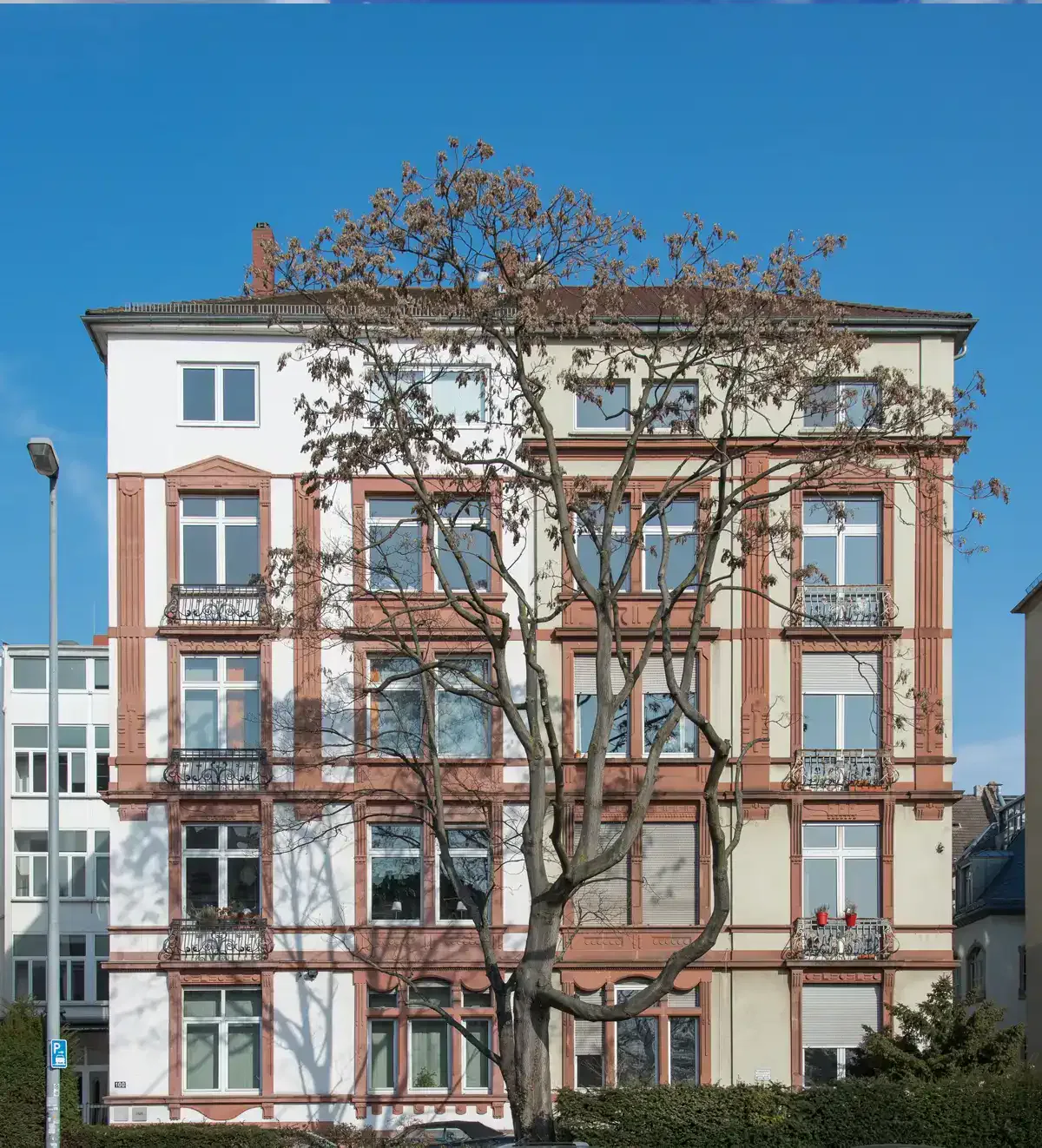 Ein vierstöckiges Wohngebäude im Gutleutviertel mit einer weiß-beigen Fassade mit roten Akzenten. Das Gebäude verfügt über mehrere Balkone mit verzierten Geländern. Davor wirft ein großer Baum ohne Blätter Schatten auf die Fassade, während der Himmel klar und blau bleibt.