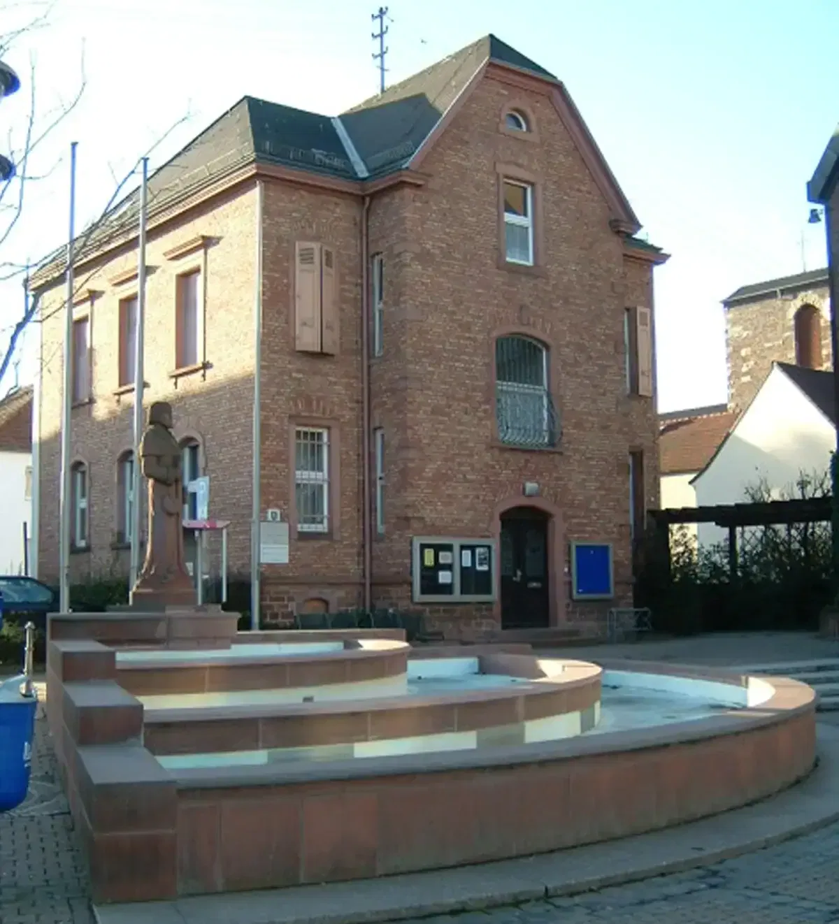 Hinter einem mehrstufigen Brunnenbau in Harheim steht ein Backsteingebäude mit Bogenfenstern. Der Brunnen ist leer, in der Mitte der untersten Stufe steht eine Statue. Der Platz ist mit Kopfsteinpflaster gepflastert, darüber ist ein klarer Himmel zu sehen.