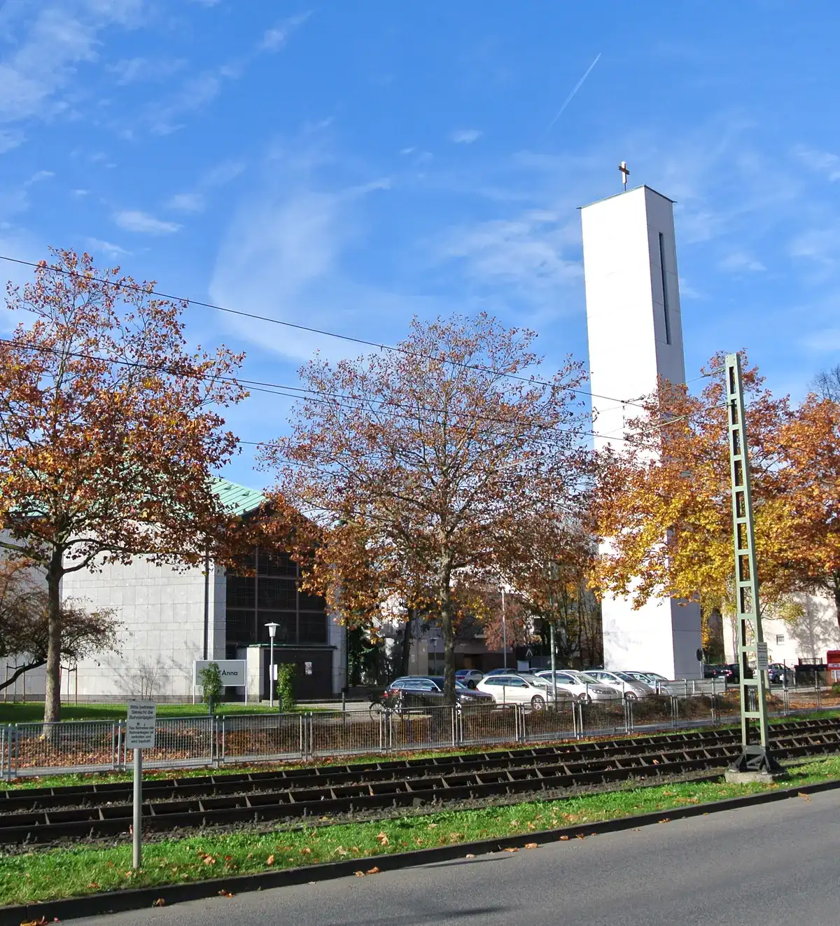 An einem klaren, sonnigen Tag stehen hinter den Gleisen in Hausen Holzbäume mit Herbstlaub, ein weißer Turm mit einem Kreuz darauf und ein modernes Kirchengebäude mit grünem Dach. Um das Gebäude herum sind mehrere geparkte Autos zu sehen. Im Vordergrund sind eine Straße und ein Wegweiser zu sehen.