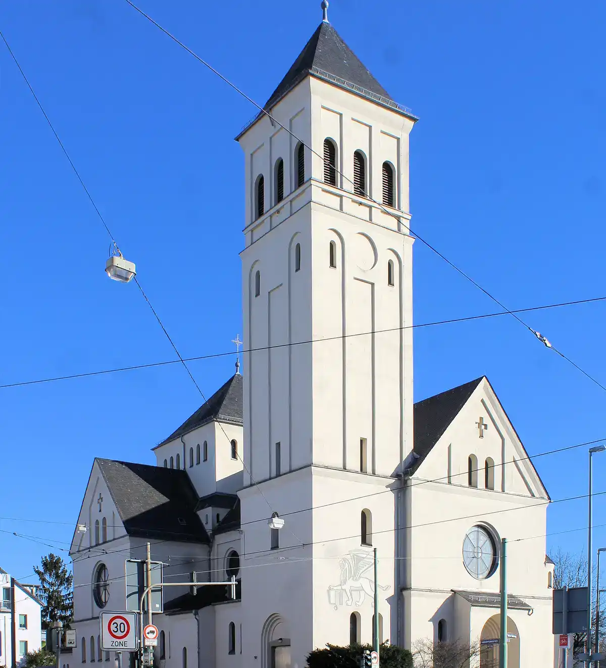 Unter einem klaren blauen Himmel im Stadtteil Nied steht eine hohe, weiße Kirche mit einem großen Kirchturm und zahlreichen Fenstern. Das Gebäude verfügt über ein Satteldach und ein rundes Fenster über dem Eingang. In der Umgebung sind Stromleitungen und Straßenschilder zu sehen.