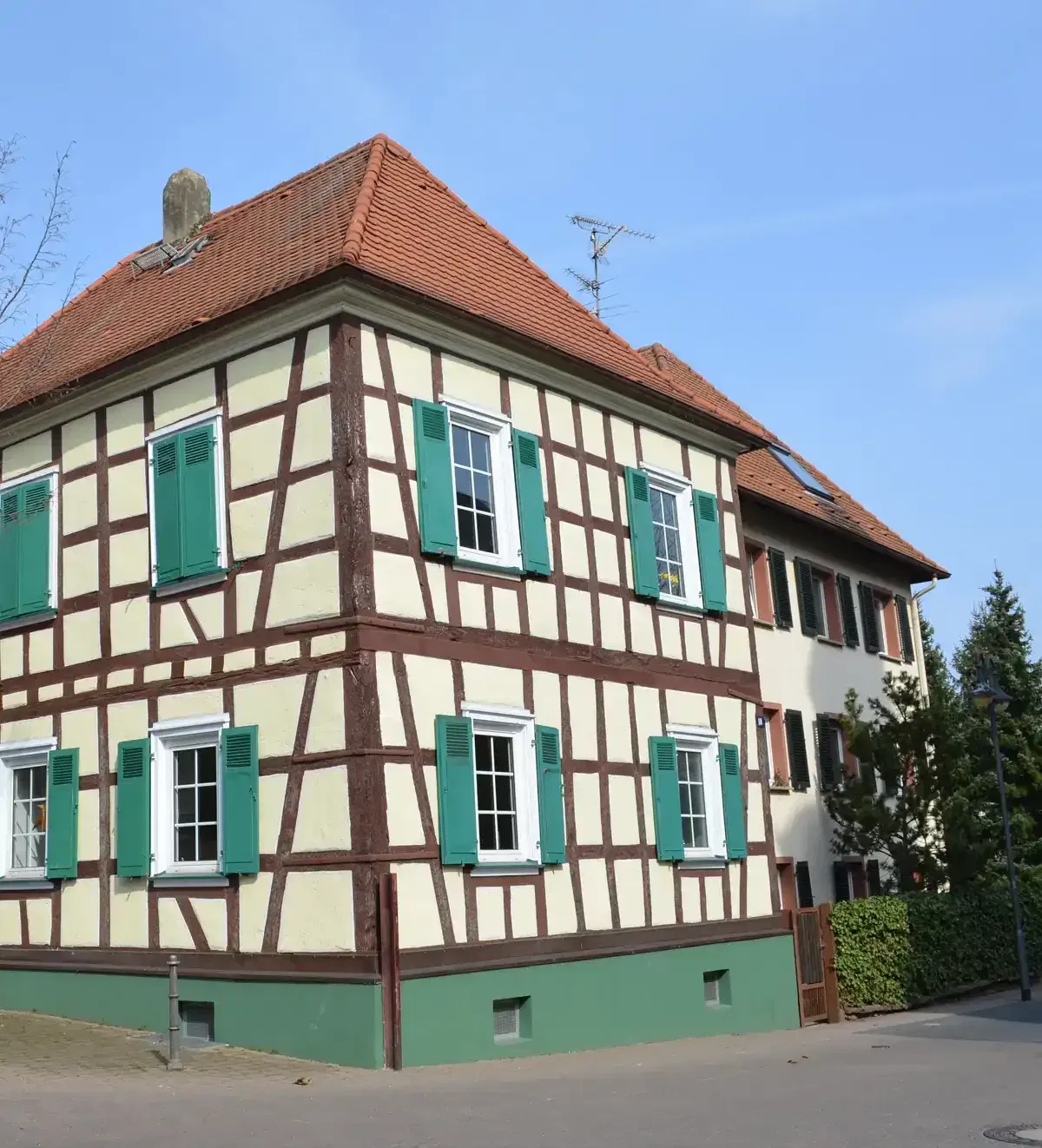 Ein traditionelles Fachwerkhaus in Nieder-Erlenbach mit weißen Wänden, dunkelbraunen Holzbalken und grünen Fensterläden. Es hat ein rotes Ziegeldach und liegt an einer gepflasterten Straße unter einem klaren blauen Himmel. Im Hintergrund ist teilweise ein weiteres Gebäude zu sehen.
