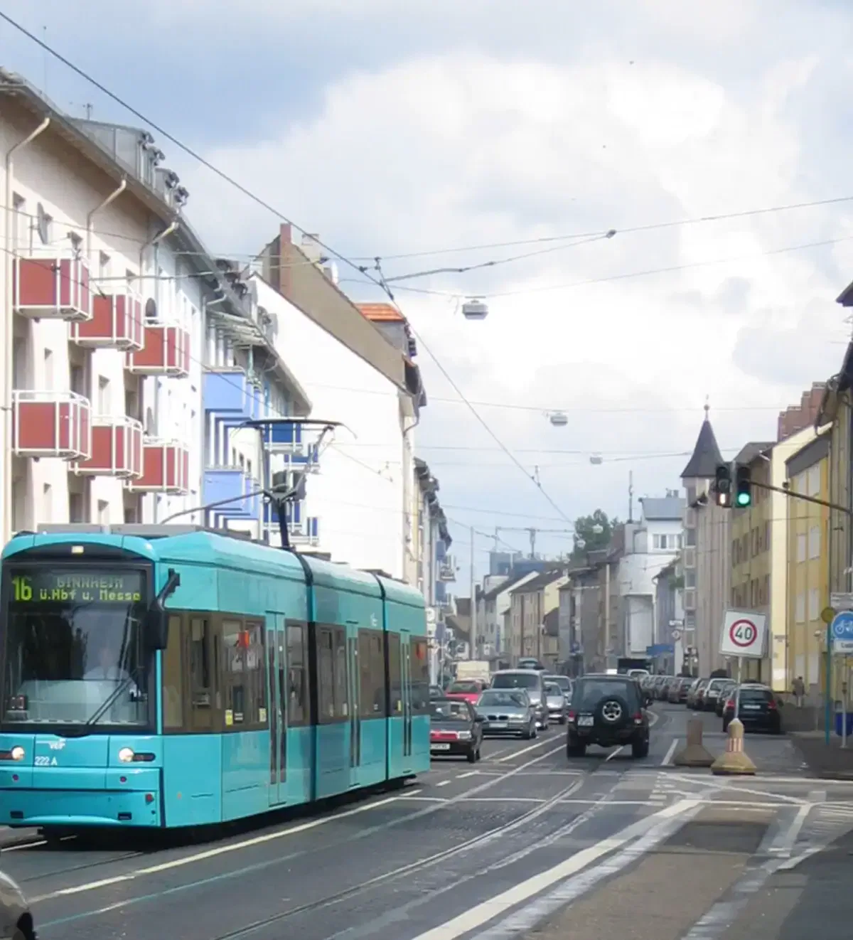 Eine blaue Straßenbahn fährt an einem bewölkten Tag eine Stadtstraße in Oberrad entlang, die von Gebäuden und Autos übersät ist. Die Straße ist von Wohn- und Geschäftshäusern gesäumt und es sind mehrere Oberleitungen und Ampeln zu sehen. Auf den Gehwegen sind einige Fußgänger zu sehen.