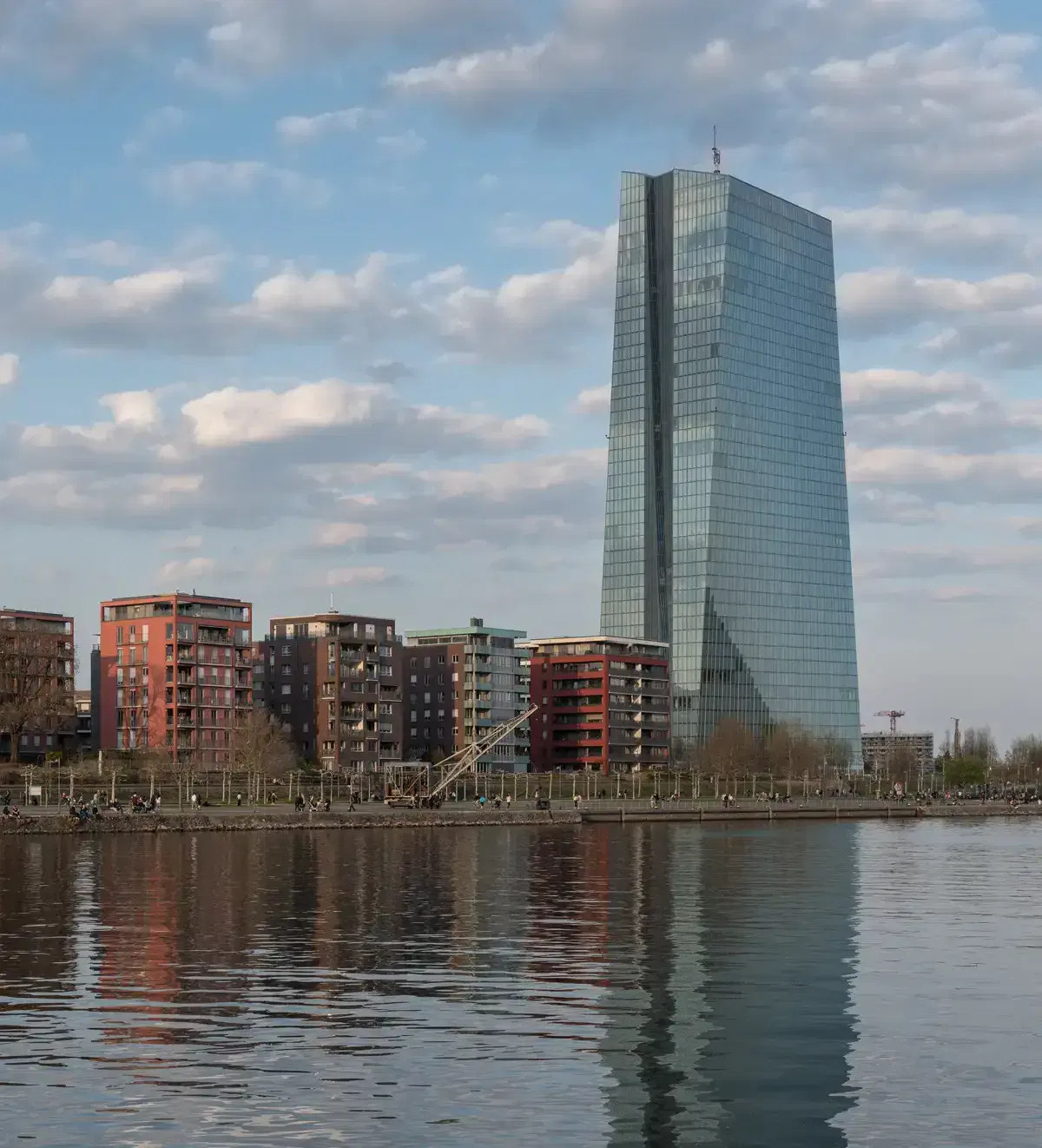 Ein hoher, moderner Wolkenkratzer aus Glas in Ostende steht neben kleineren, farbenfrohen Wohnhäusern entlang einer Uferpromenade. Der Himmel ist teilweise bewölkt und die Gebäude spiegeln sich im ruhigen Wasser darunter.
