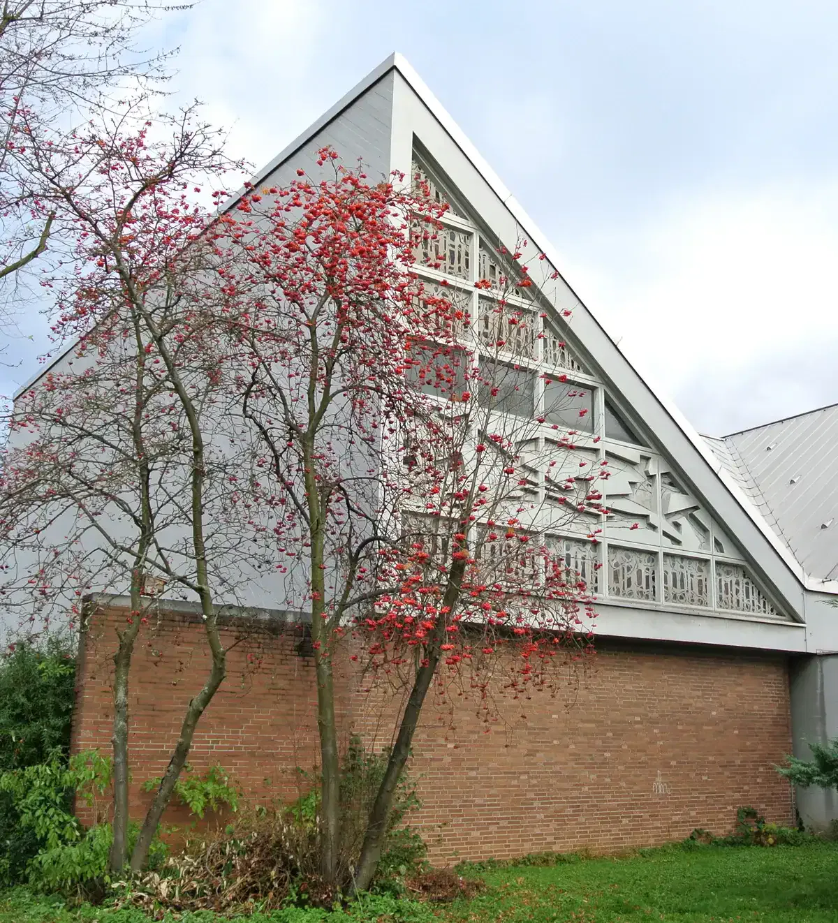 Ein dreieckiges Haus in Preungesheim mit aufwendiger weißer Holzverkleidung steht hinter einem Baum mit roten Beeren. Der untere Teil ist aus Ziegeln, und der obere Teil hat eine graue Außenfassade. Der Himmel ist bedeckt und am Fuß des Baumes ist etwas Grün zu sehen.