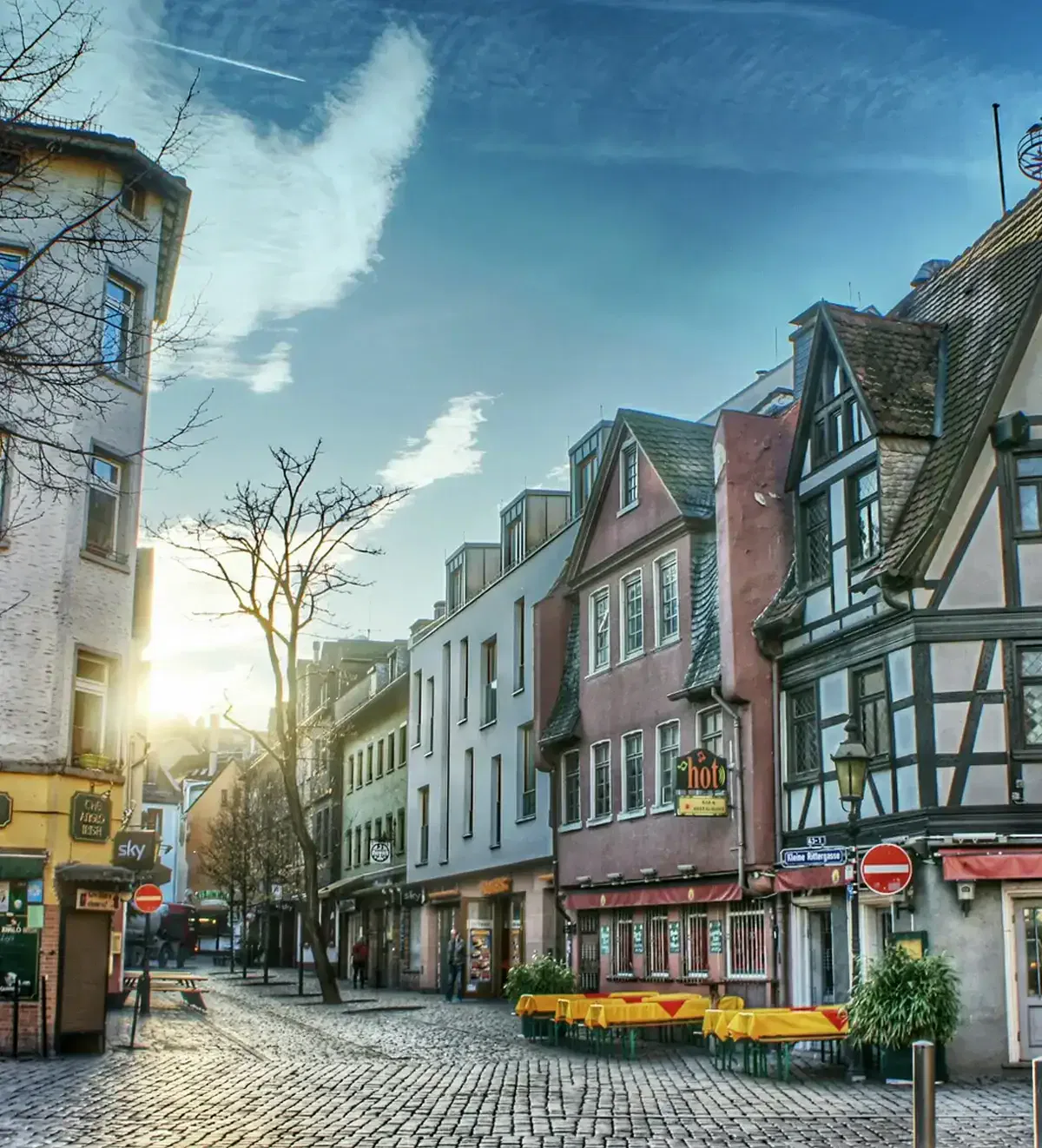 frankfurt-stadtteile-sachsenhausen Eine malerische Straßenszene in der alten europäischen Stadt Sachsenhausen zeichnet sich durch Fachwerkhäuser und Kopfsteinpflasterstraßen aus. Im Hintergrund geht die Sonne unter und wirft einen warmen Schein auf die Straße. Sitzplätze im Freien säumen den Bürgersteig, und ein paar Bäume verleihen der Straße Charme.