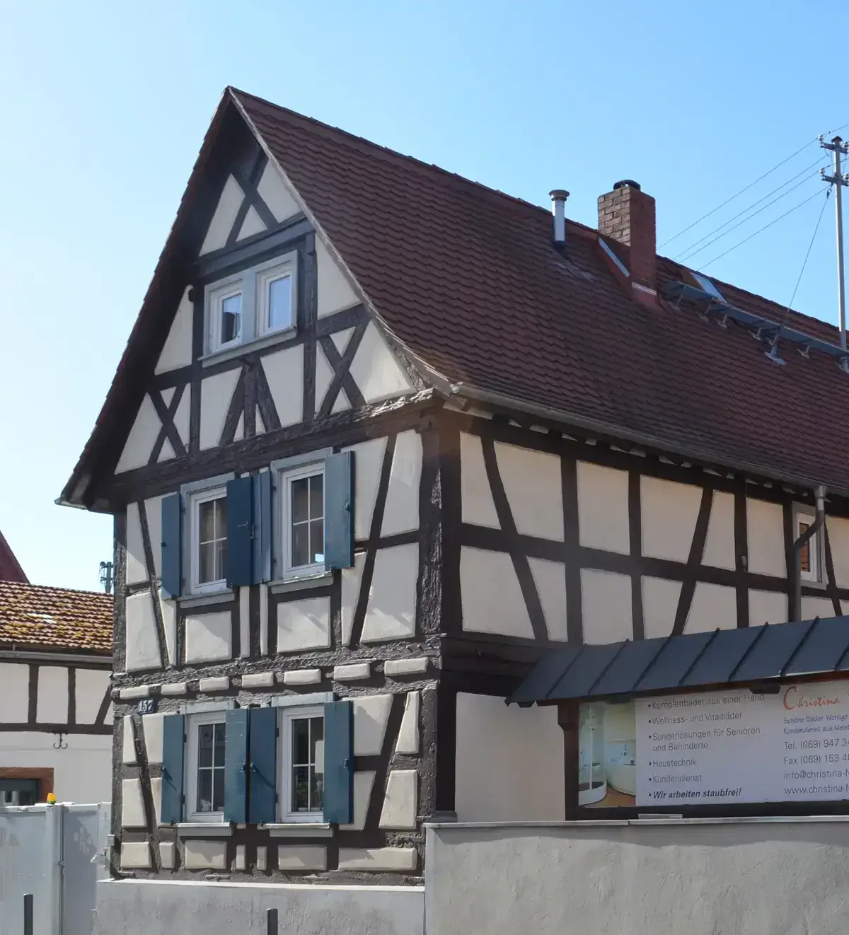 Ein traditionelles Fachwerkhaus in Seckbach zeichnet sich durch ein steiles Satteldach, blaue Fensterläden und weißen Putz zwischen dunklen Holzbalken aus. Auf dem Dach befindet sich ein kleines Dachfenster und über einer Tür ragt ein Vordach hervor. Ein Schild auf der rechten Seite bietet Geschäftsinformationen.