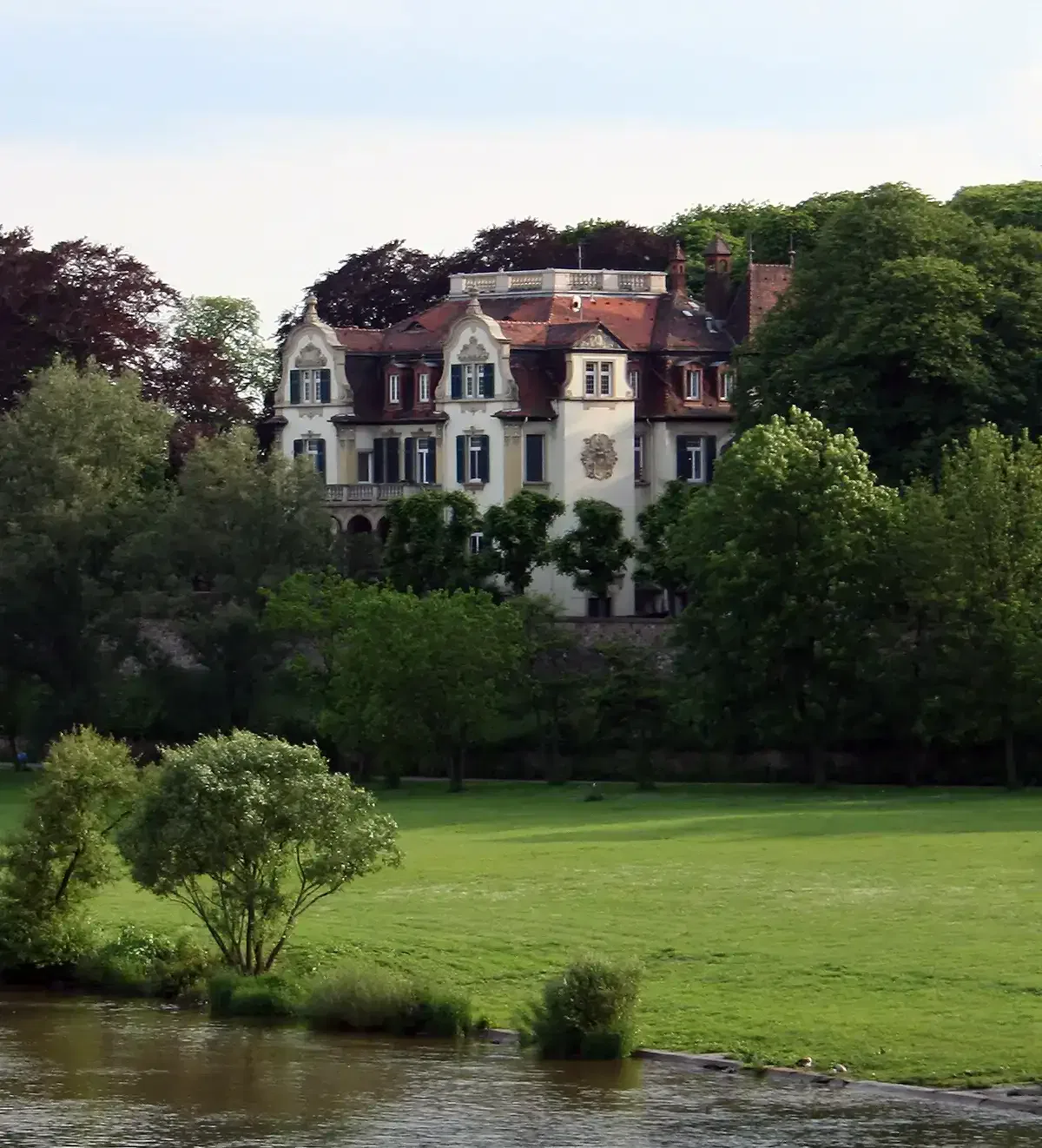 frankfurt-stadtteile-sindlingen Ein großes, elegantes Herrenhaus mit kunstvollen architektonischen Details wird teilweise von üppigen grünen Bäumen verdeckt. Das Gebäude liegt eingebettet in Sindlingen, ist von einem weitläufigen grünen Rasen umgeben und liegt in der Nähe eines ruhigen Flusses, was eine heitere und malerische Szenerie schafft.