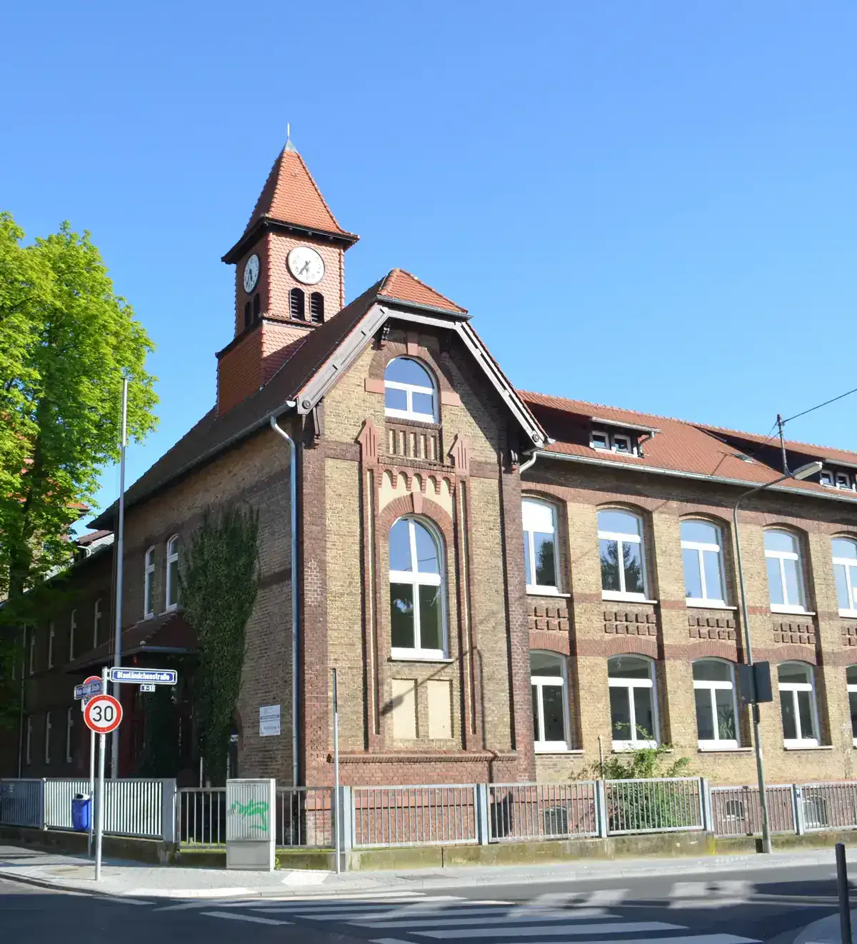 Ein historisches Backsteingebäude mit rotem Ziegeldach und Uhrturm steht stolz unter einem klaren blauen Himmel in Zeilsheim. Das Gebäude verfügt über große Bogenfenster im zweiten Stock und rechteckige Fenster im ersten Stock. Vorne weist ein Straßenschild auf eine Geschwindigkeitsbegrenzung von 30 km/h hin.