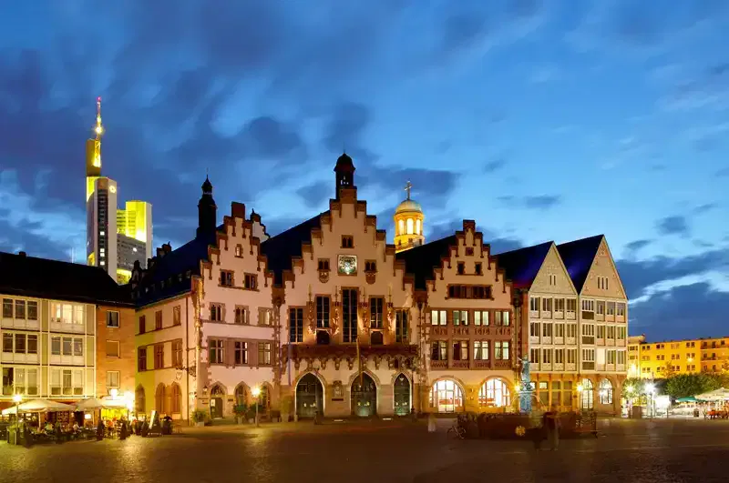 Eine malerische Abendansicht des Römergebäudes in Frankfurt, Deutschland, beleuchtet unter einem Abendhimmel. Das historische Gebäude mit seiner markanten Giebelfassade steht prominent auf dem Römerberg neben dem Gerechtigkeitsbrunnen, umgeben von modernen Wolkenkratzern und anderen Gebäuden.