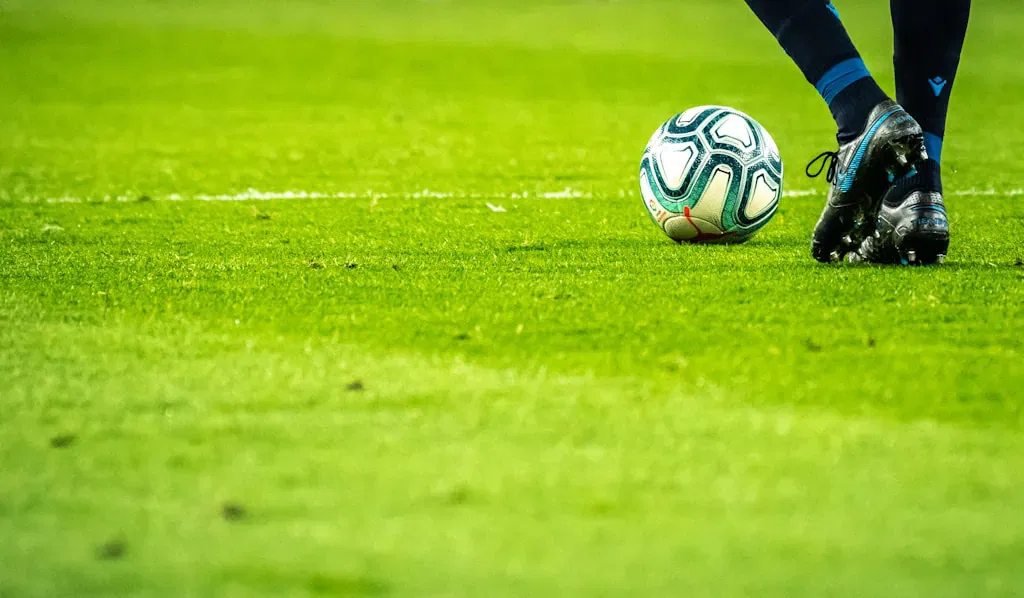 Eine Nahaufnahme der Beine und Schuhe eines Fußballspielers, der sich darauf vorbereitet, einen Fußball auf einem grünen Rasenfeld zu kicken; ein automatisch gespeicherter Entwurf fängt die dunklen Socken und Stollenschuhe perfekt ein.